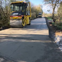 Żółty walec Dynapac CC232HF ubija świeżo wylaną nawierzchnię drogi; w tle drugi walec i pracownik w odblaskowej kamizelce.