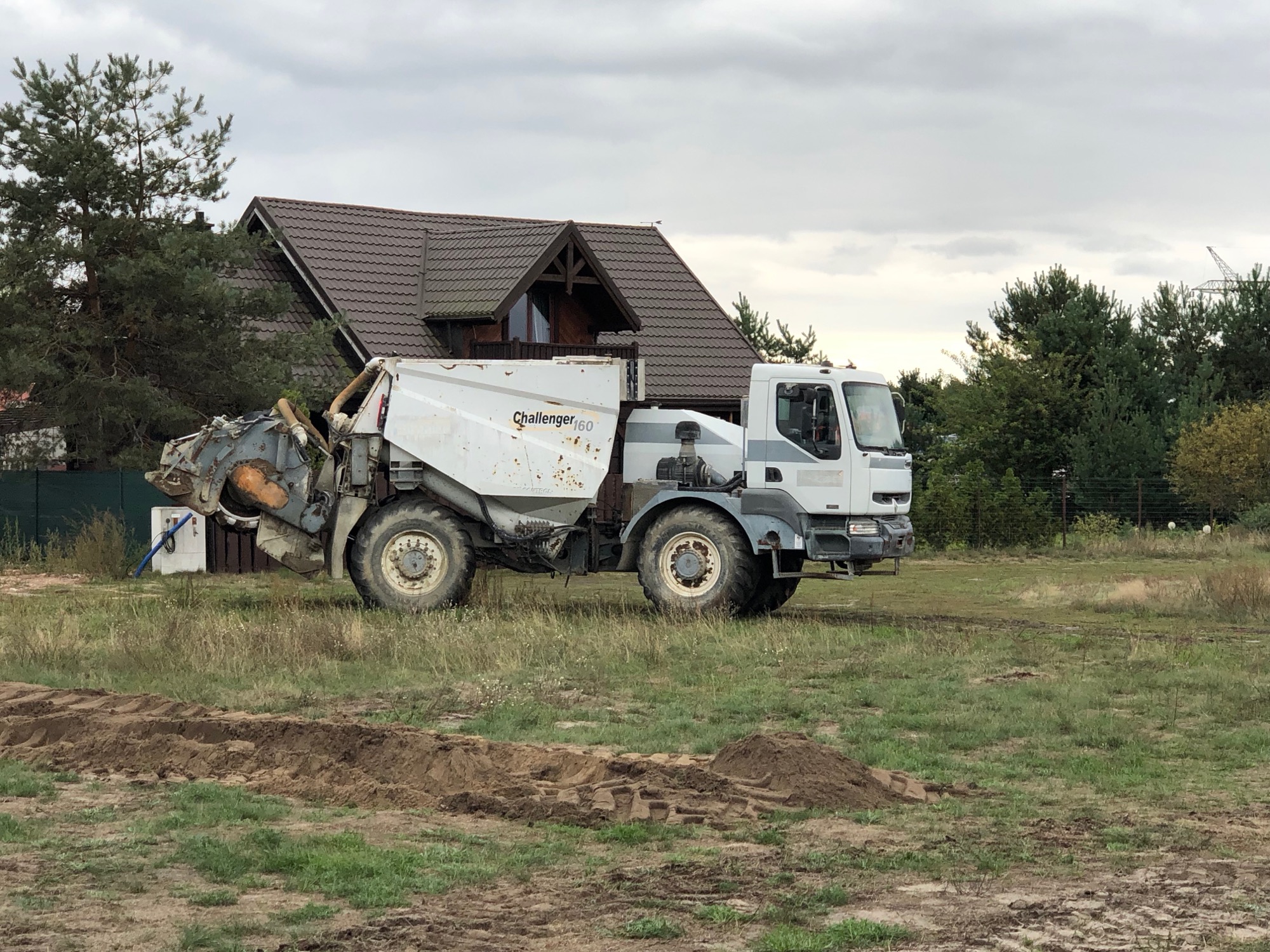 Specjalistyczna maszyna Challenger 160, prawdopodobnie frezarka do pni, stoi na trawiastym terenie z widocznymi śladami prac ziemnych, w tle dom z brązowym dachem i drzewa pod pochmurnym niebem.