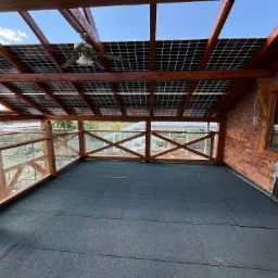 Zadaszony taras z panelami słonecznymi, drewnianą konstrukcją i balustradą ze szkła. Widoczna czarna, płaska powierzchnia dachu i ceglana ściana budynku.