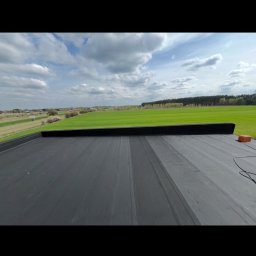 Hydroizolacje Makowski Konrad - Widok z góry na płaski, czarny dach z papy, z widocznym fragmentem uszczelnienia i zielonym krajobrazem w tle, pod błękitnym niebem z chmurami.