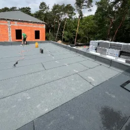 Montaż papy termozgrzewalnej na płaskim dachu budynku w stanie surowym otwartym, widoczne narzędzia i materiały izolacyjne.