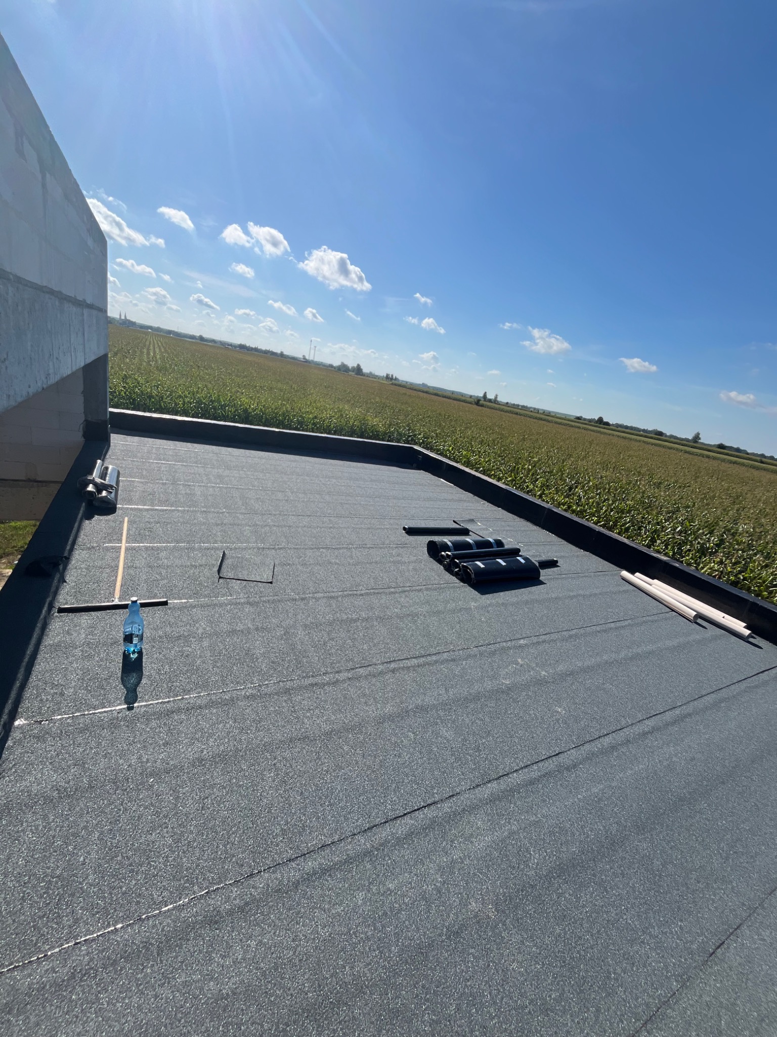 Gotowy, płaski dach z papy termozgrzewalnej na budynku w budowie, z widocznymi narzędziami i materiałami. W tle pole uprawne pod błękitnym niebem.