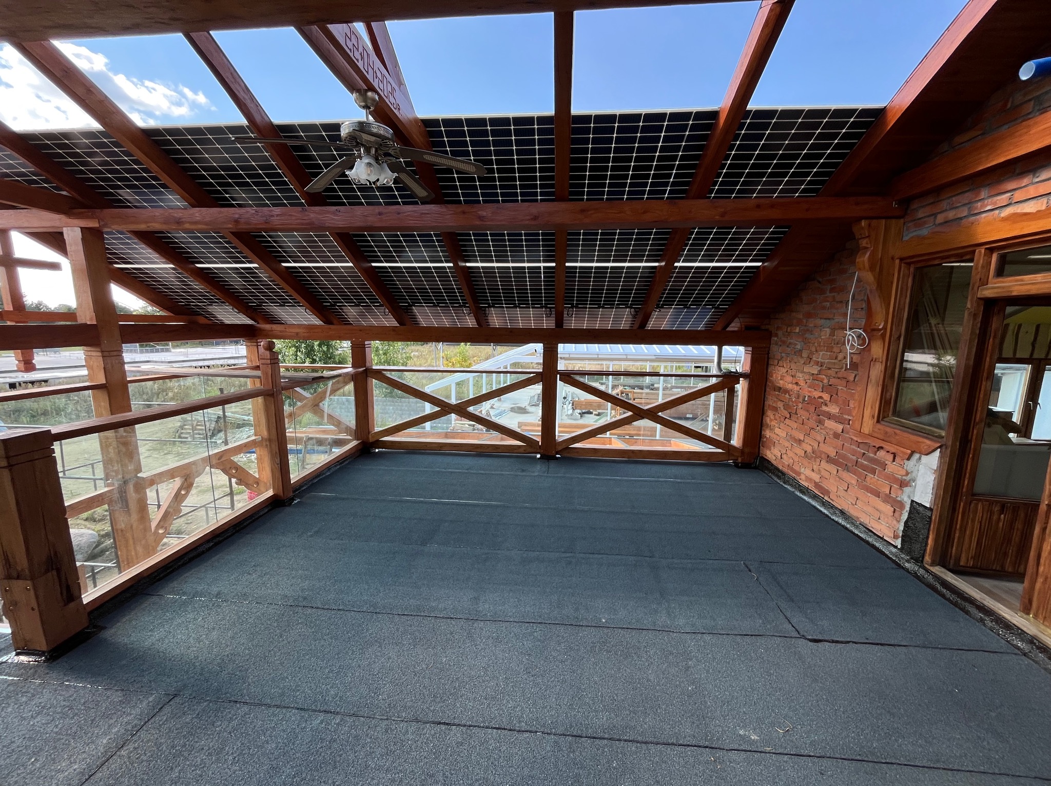 Zadaszony taras z panelami słonecznymi, drewnianą konstrukcją i balustradą ze szkła. Widoczna czarna, płaska powierzchnia dachu i ceglana ściana budynku.