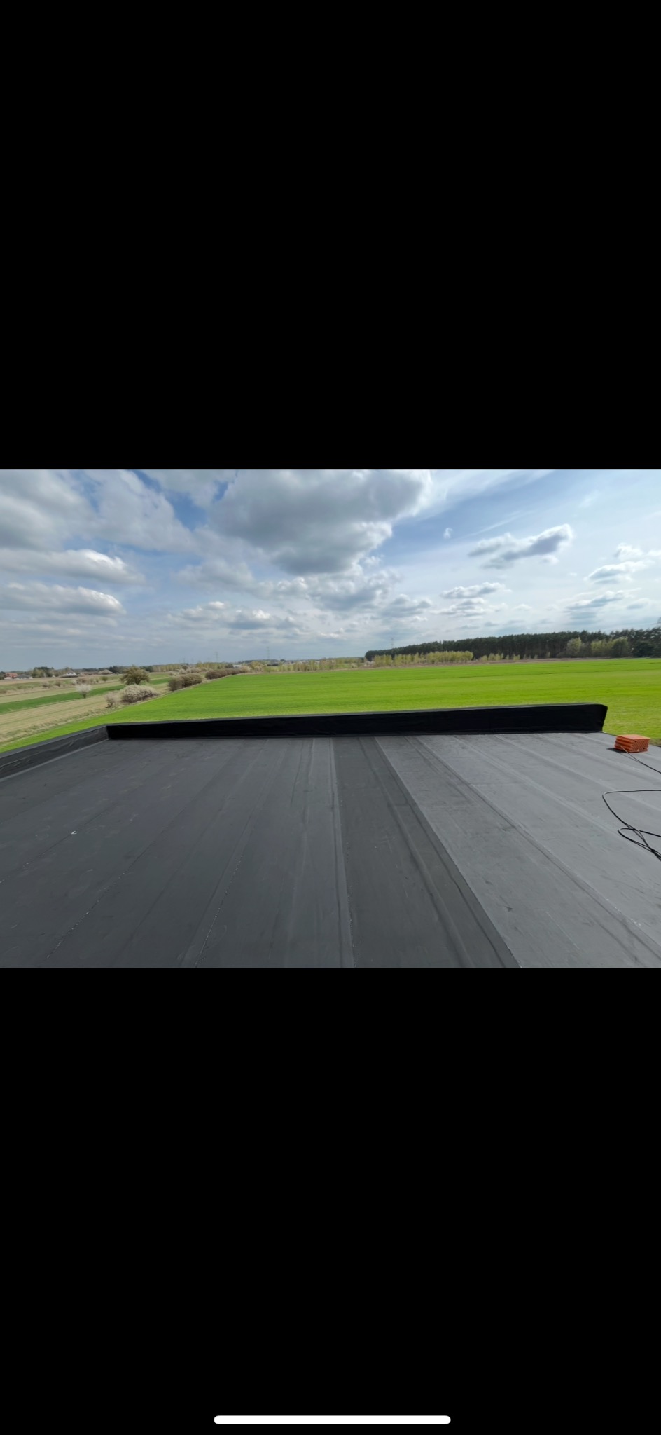 Widok z góry na płaski, czarny dach z papy, z widocznym fragmentem uszczelnienia i zielonym krajobrazem w tle, pod błękitnym niebem z chmurami.