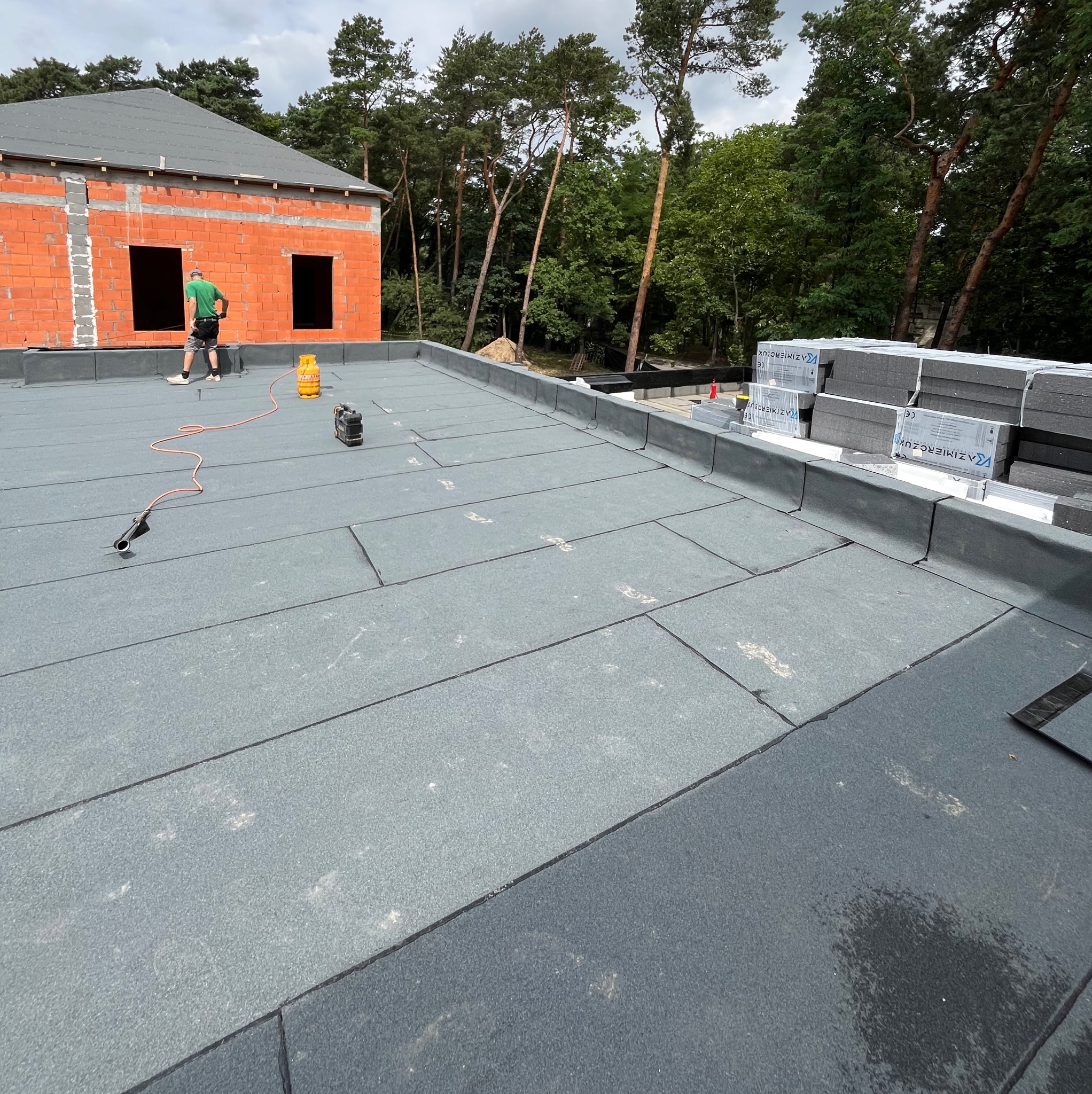 Montaż papy termozgrzewalnej na płaskim dachu budynku w stanie surowym otwartym, widoczne narzędzia i materiały izolacyjne.