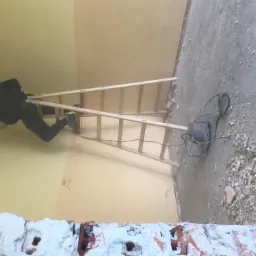 Pracownik na drabinie szpachluje ścianę w pomieszczeniu w trakcie remontu, widoczne gruz i pył na podłodze, ślady po kuciu na ścianie.