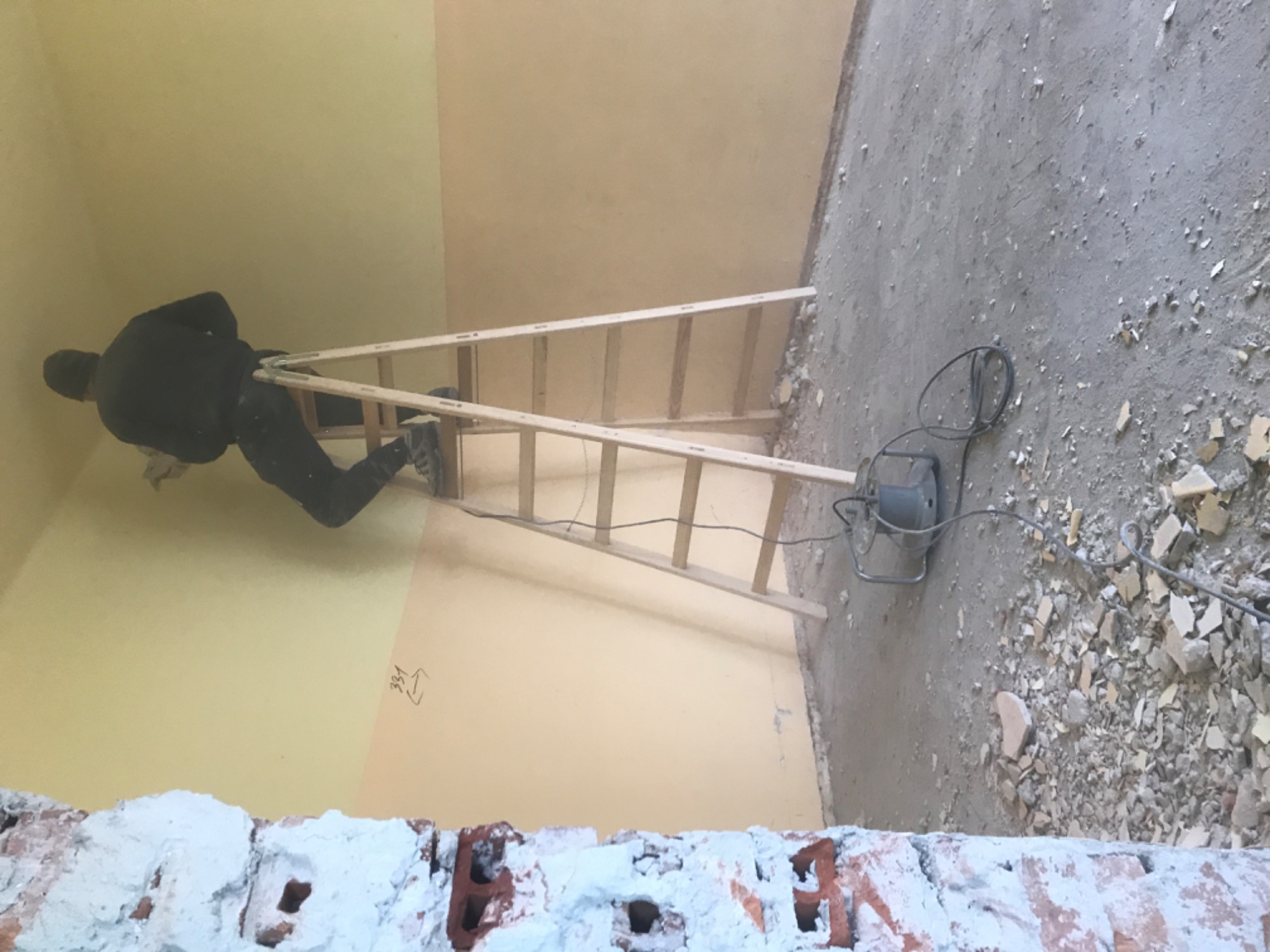 Pracownik na drabinie szpachluje ścianę w pomieszczeniu w trakcie remontu, widoczne gruz i pył na podłodze, ślady po kuciu na ścianie.