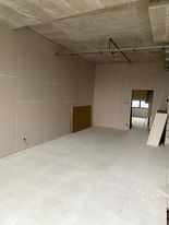 Wnętrze w trakcie remontu: surowe ściany pokryte gładzią, widoczne płyty na podłodze i surowy betonowy sufit z instalacjami. Otwór drzwiowy prowadzi do jaśniejszego pomieszczenia.