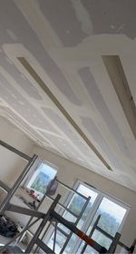 Montaż płyty karton gips na suficie, widoczne profile i szpachlowane łączenia, rusztowanie w pomieszczeniu z oknem.
