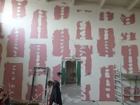 Ściana w trakcie szpachlowania, widoczne różowe plamy gipsu na białej powierzchni, przygotowanie pod malowanie, metalowa drabina oparta o ścianę, widoczne drzwi w tle.