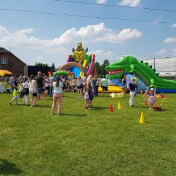Plenerowa impreza z dmuchanym placem zabaw w kształcie krokodyla i zjeżdżalnią z motywem Minionka, otoczona przez uczestników, na tle zielonego trawnika i budynków.