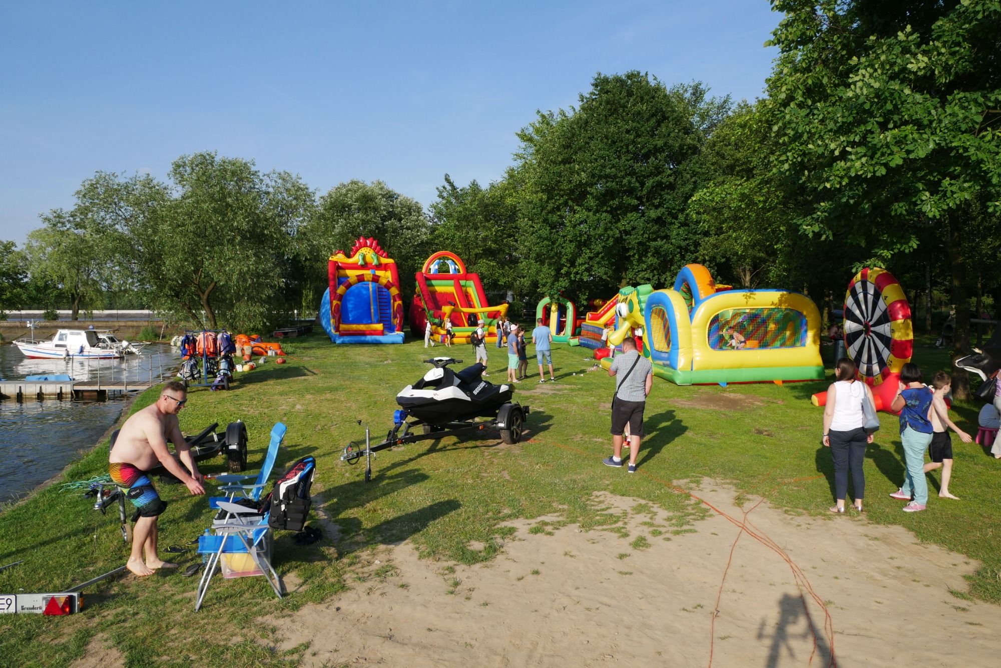 Rozstawione dmuchane zamki i zjeżdżalnie dla dzieci na trawiastym terenie nad wodą, z widocznym skuterem wodnym na przyczepce i osobami w tle.