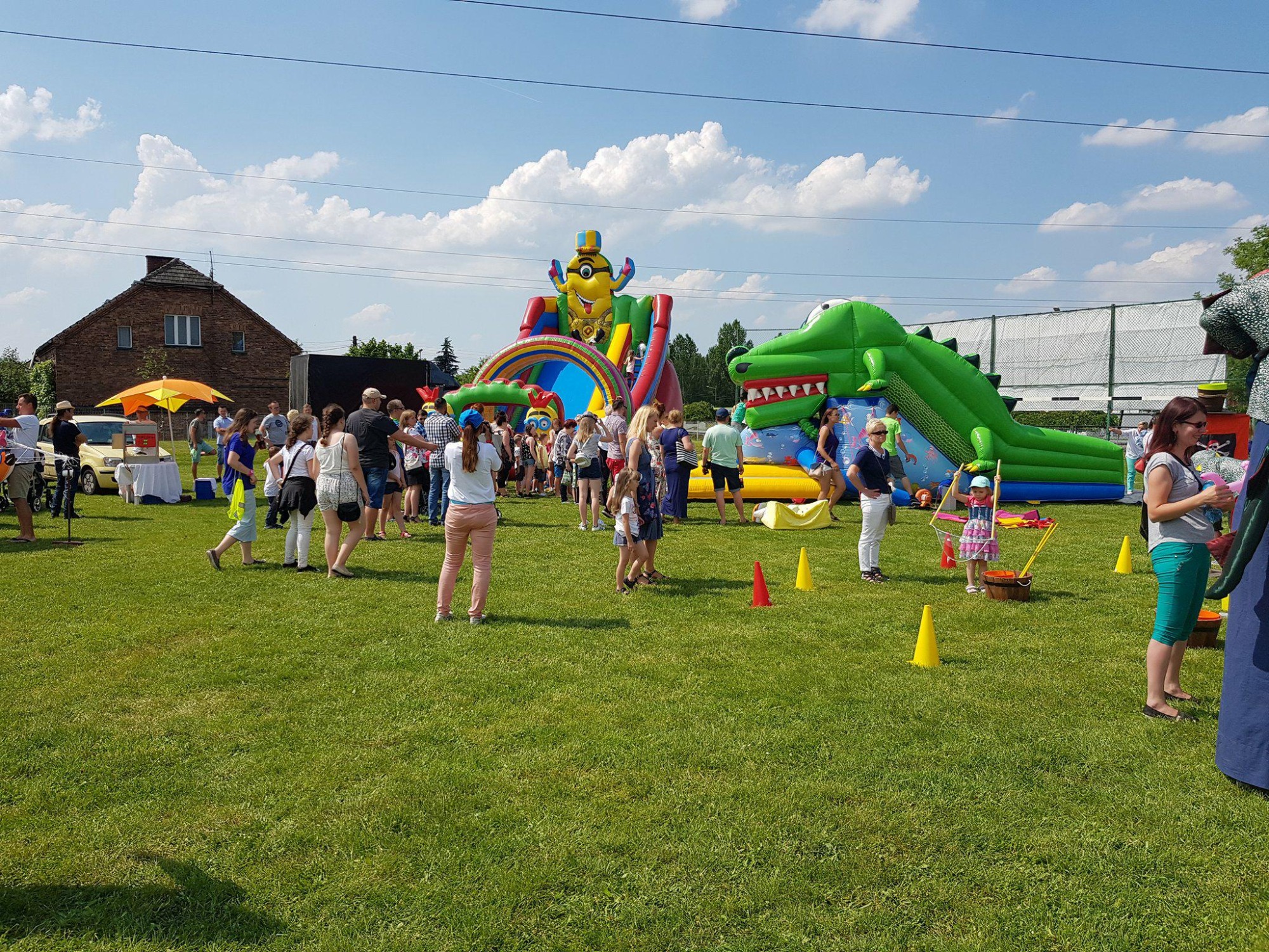 Plenerowa impreza z dmuchanym placem zabaw w kształcie krokodyla i zjeżdżalnią z motywem Minionka, otoczona przez uczestników, na tle zielonego trawnika i budynków.
