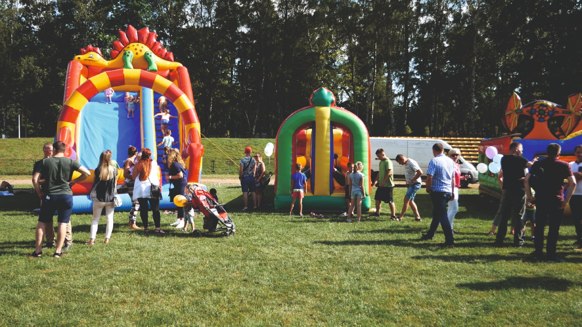 Kolorowe dmuchańce, w tym zjeżdżalnia w kształcie dinozaura i tunel, na zielonym trawniku w Grojcu, otoczone grupą ludzi, dzieci bawiące się na dmuchańcach.