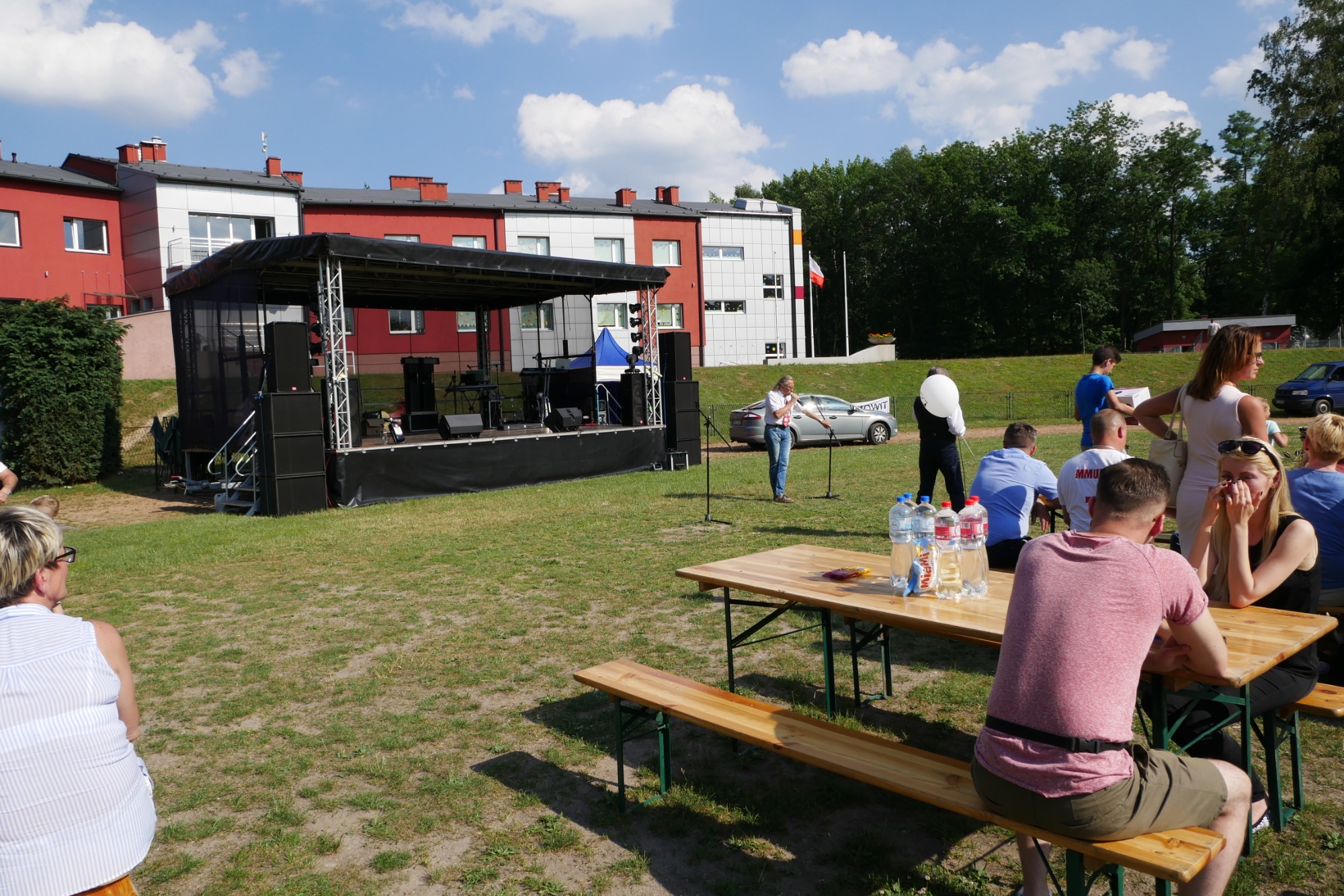 Scena plenerowa z nagłośnieniem i oświetleniem, przygotowana do wydarzenia na trawiastym terenie, z widocznymi stolikami i siedzącymi osobami.