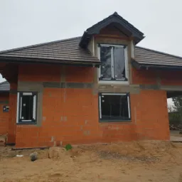 Surowy stan domu w trakcie budowy z zamontowanymi oknami z czarnymi ramami, widoczne bloczki z czerwonej cegły i brązowa dachówka, teren wokół domu zasypany piaskiem.