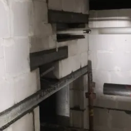 Wznoszenie ściany z białych bloczków, wzmocnionej metalowymi profilami, z widocznymi otworami na instalacje i wspornikami rusztowania.