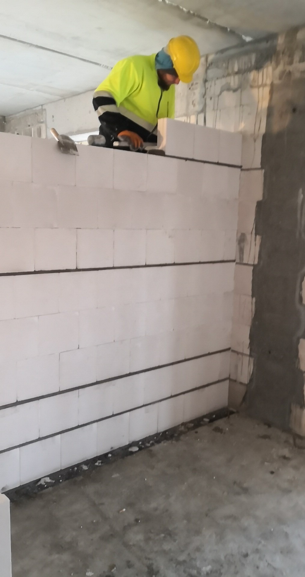 Murarz w żółtym kasku i odblaskowej kamizelce układa białe bloczki z betonu komórkowego, widoczna kielnia i zaprawa, wnętrze surowego budynku w trakcie budowy.