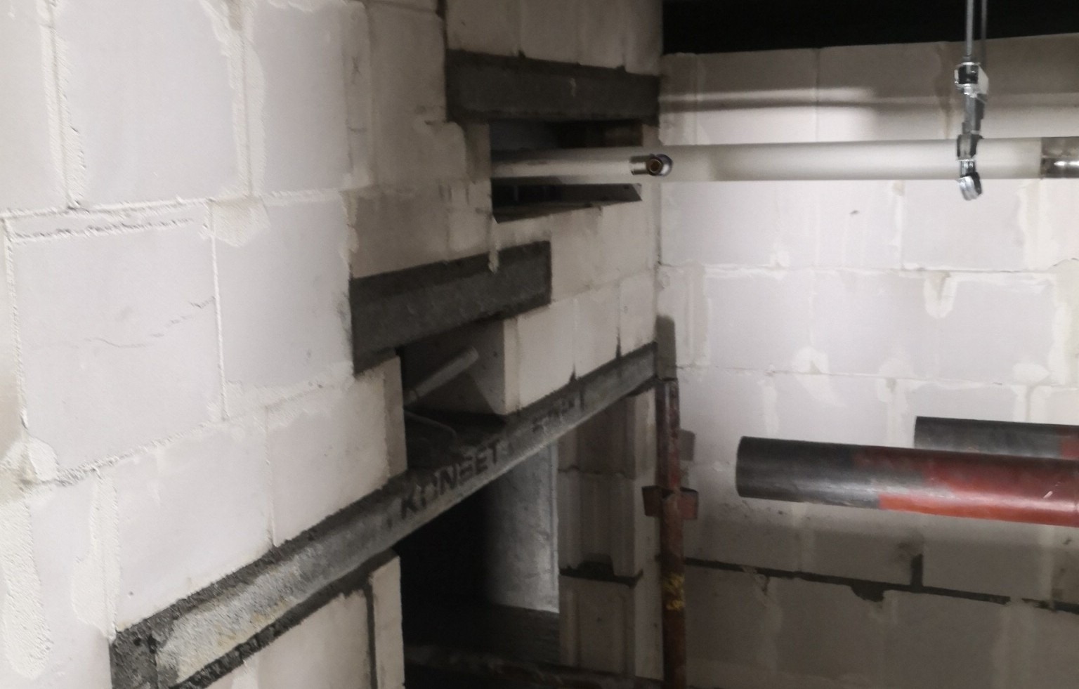 Wznoszenie ściany z białych bloczków, wzmocnionej metalowymi profilami, z widocznymi otworami na instalacje i wspornikami rusztowania.