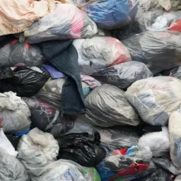 Stos worków z odzieżą używaną, widoczne dżinsy na wierzchu, różne kolory i materiały ubrań widoczne przez przezroczyste worki.