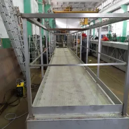 Metalowa konstrukcja platformy roboczej z kratownicą, widoczny spawacz w czerwonej czapce w hali produkcyjnej, drabina oparta o ścianę w tle.