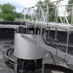 Metalowa konstrukcja i zbiornik w oczyszczalni ścieków widziane z góry, z widoczną metalową kładką i zielenią w tle.