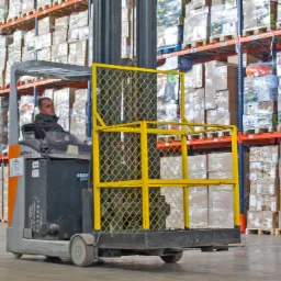 Operator w wózku widłowym STILL z żółtą platformą roboczą na tle regałów magazynowych wypełnionych kartonami i opakowaniami owiniętymi folią.