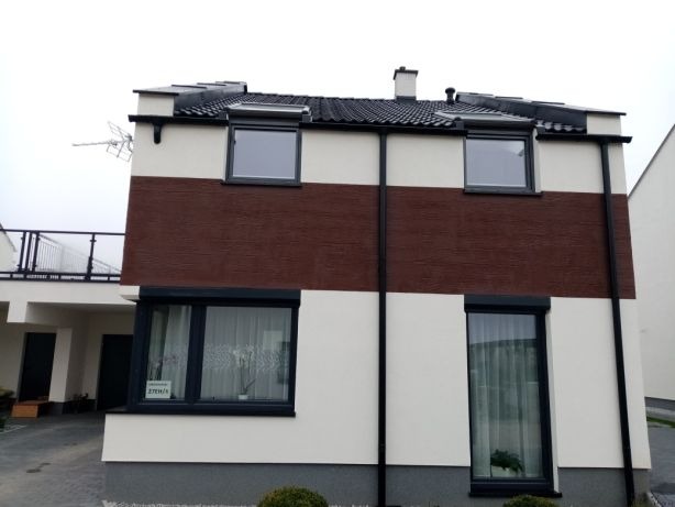 Dwukondygnacyjny dom z elewacją w pasy: biały tynk i brązowa struktura, czarne rynny i okna z roletami zewnętrznymi, widok z zewnątrz.