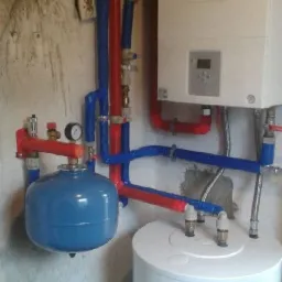 Instalacja kotła gazowego z niebieskim zbiornikiem wyrównawczym, widoczne rury w kolorze czerwonym i niebieskim oraz biały zasobnik ciepłej wody, zamontowane na tle surowej ściany.