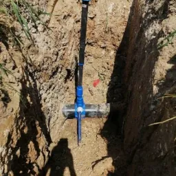 Wykop z widocznym przyłączem wodociągowym z rurą poziomą i pionowym wyprowadzeniem zakończonym białym korkiem, cień osoby fotografującej na dnie wykopu.