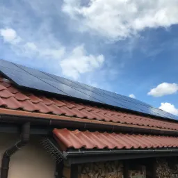 Czerwona dachówka z zainstalowanymi panelami słonecznymi w słoneczny dzień z widocznymi chmurami na niebie.