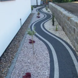 Kręty chodnik z kostki brukowej z białymi pasami, obsypany drobnym kamieniem, z młodymi drzewkami i niską ścianą z kamienia w tle.