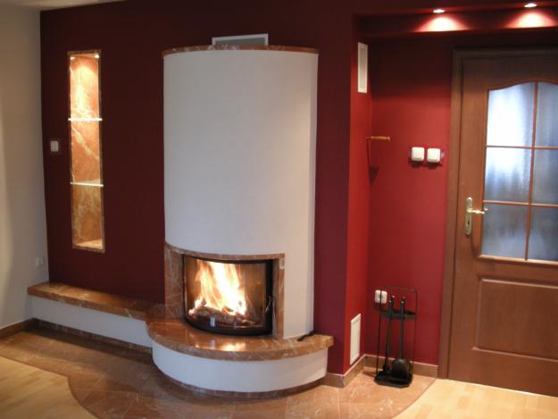 Nowoczesny kominek z obudową z jasnego marmuru i białym korpusem, płomienie w palenisku, w tle ściana w kolorze bordo z wnęką wykończoną marmurem.