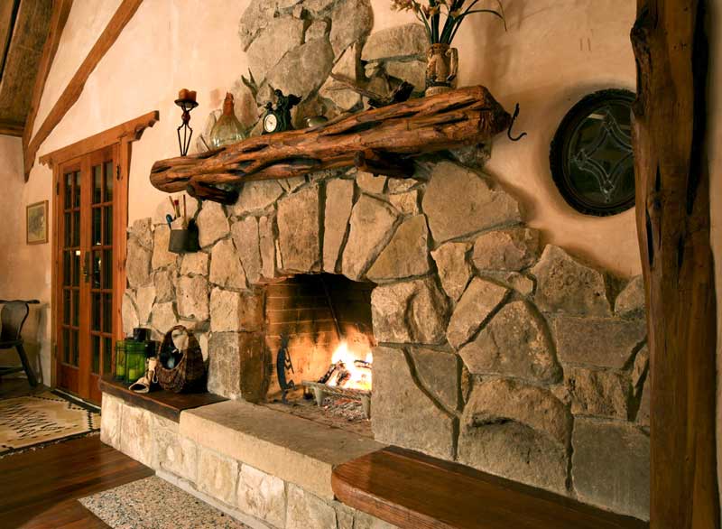 Rustykalny kominek z kamienia z paleniskiem, nad którym znajduje się półka z pnia drzewa, a na niej dekoracje: świecznik, zegar i wazon z kwiatami.