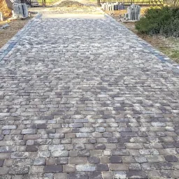 Ułożona ścieżka z kostki brukowej w odcieniach szarości i brązu, z ciemniejszym obramowaniem, prowadząca przez posesję, z widocznymi stosami kostki do dalszych prac.