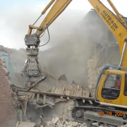 Żółta koparka gąsienicowa Auto-Box z hydraulicznymi nożycami do betonu rozbiera ceglany budynek, unoszący się pył i gruz.