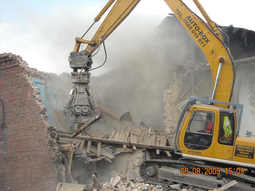 Żółta koparka gąsienicowa Auto-Box z hydraulicznymi nożycami do betonu rozbiera ceglany budynek, unoszący się pył i gruz.