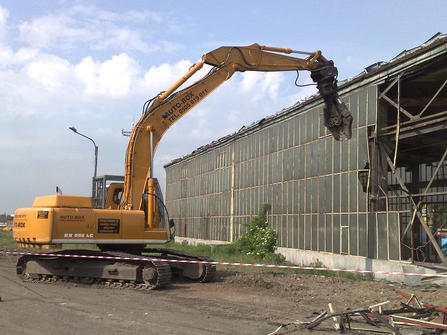 Żółta koparka gąsienicowa z osprzętem do kruszenia, podczas pracy przy rozbiórce hali z przeszkloną ścianą, widoczne gruz i taśma ostrzegawcza.