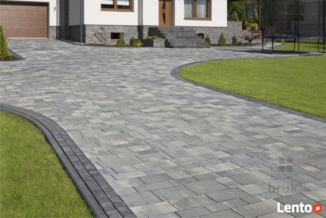 Ułożona szara kostka brukowa na podjeździe do domu z brązową bramą garażową, widoczny trawnik i trampolina w tle.