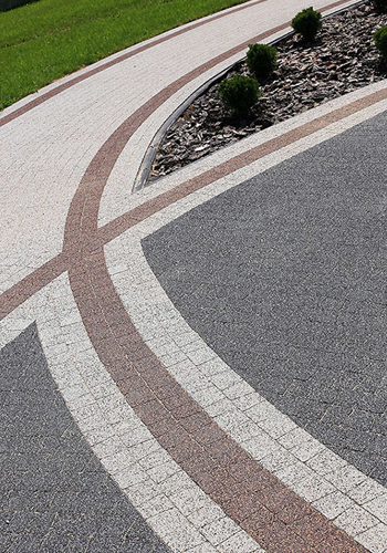 Ścieżka z kostki brukowej w odcieniach szarości i brązu, zakrzywiona linia oddzielająca kostkę od trawnika, ozdobne krzewy w tle.