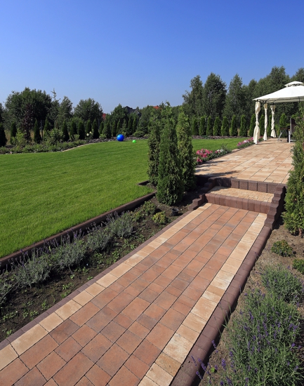 Ogród z ścieżką z kostki w odcieniach brązu i beżu, prowadzącą do altany z białymi zasłonami, na trawniku widoczne dwie kolorowe piłki.