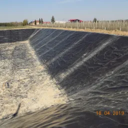 Wykopany dół wyłożony czarną folią, prawdopodobnie przygotowywany pod budowę oczka wodnego lub basenu, z widocznymi śladami piasku i ziemi wewnątrz, w tle zabudowania gospodarcze i drzewa.