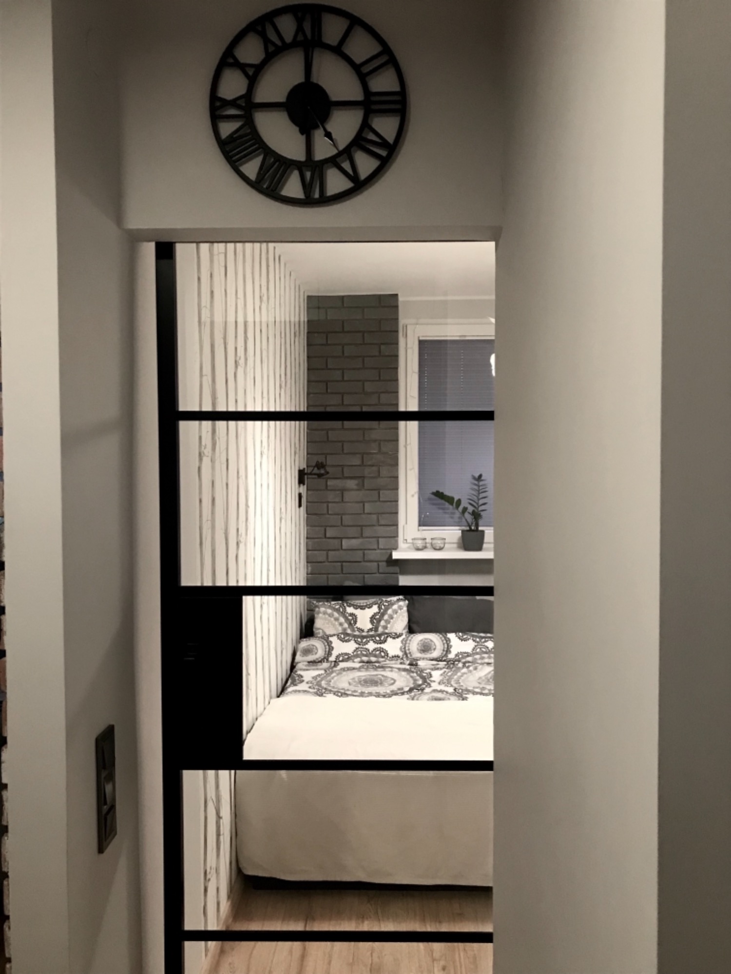 Sypialnia widziana przez szklane drzwi z czarnymi szprosami, nad drzwiami duży zegar rzymski, w pokoju łóżko z wzorzystą pościelą, ściana z cegły i okno z roletami.