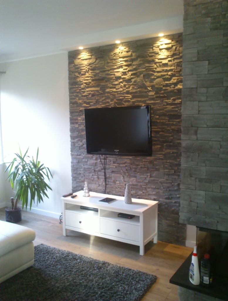 Ściana z kamienia dekoracyjnego podświetlona punktowo, z telewizorem i białą szafką RTV w salonie z drewnianą podłogą i rośliną doniczkową.