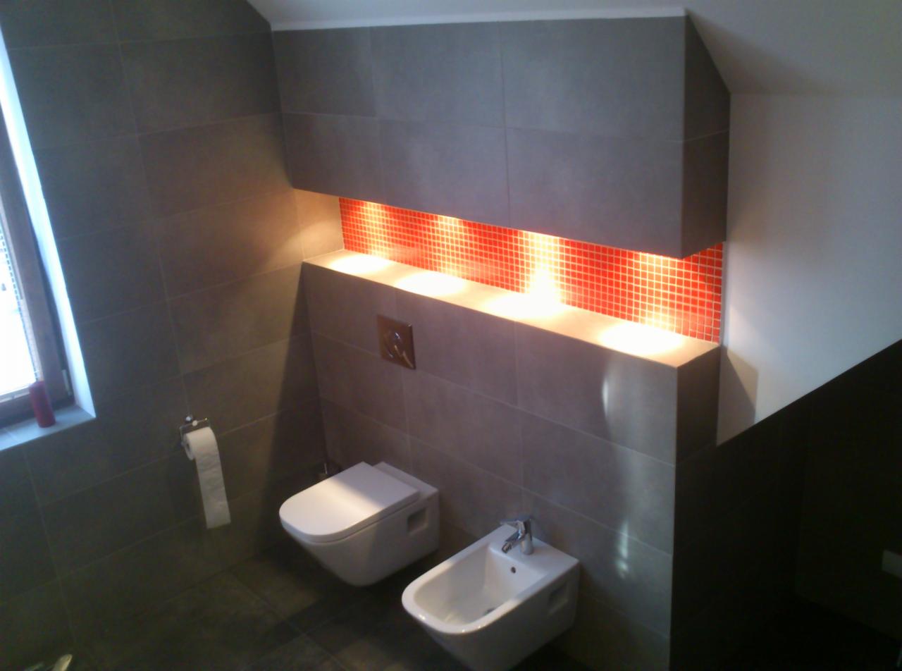 Nowoczesna łazienka z szarymi płytkami, podświetlaną wnęką z czerwoną mozaiką, wiszącą toaletą i bidetem. Ujęcie z góry, widoczne okno.