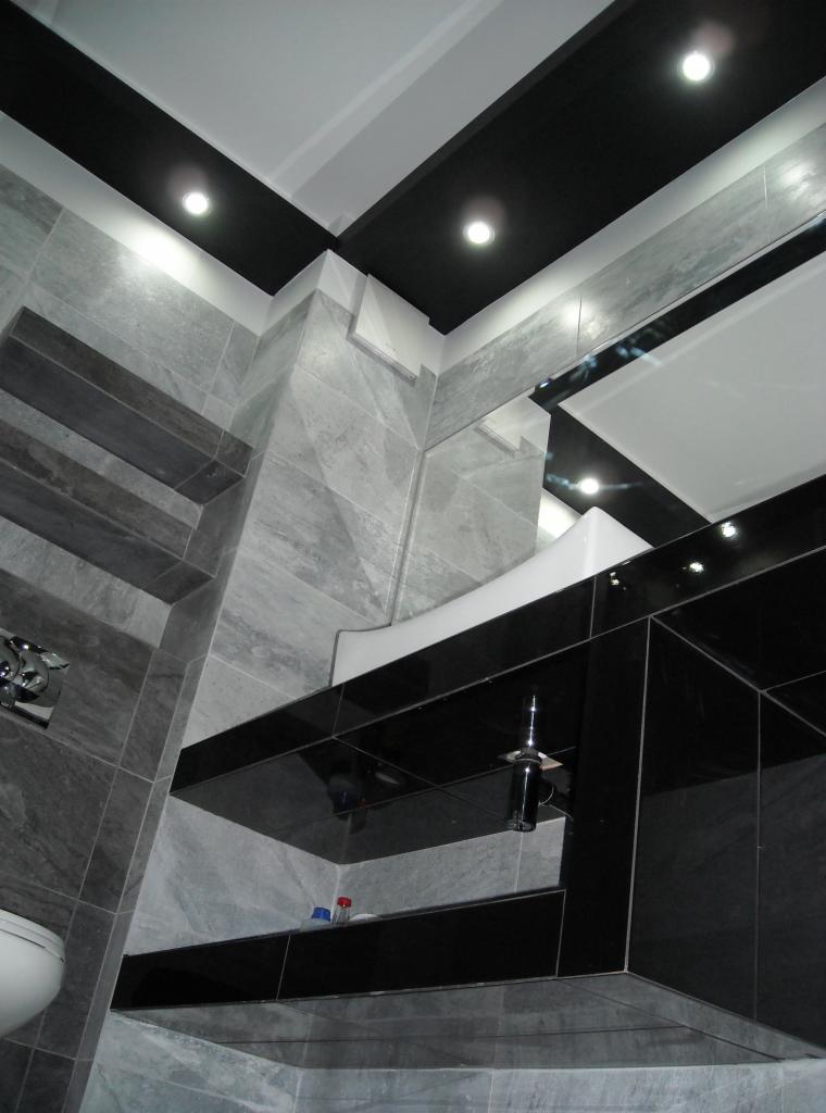 Nowoczesna łazienka z szarymi płytkami i czarnymi akcentami, oświetleniem punktowym i minimalistyczną umywalką, prezentująca wykończenie wnętrza.