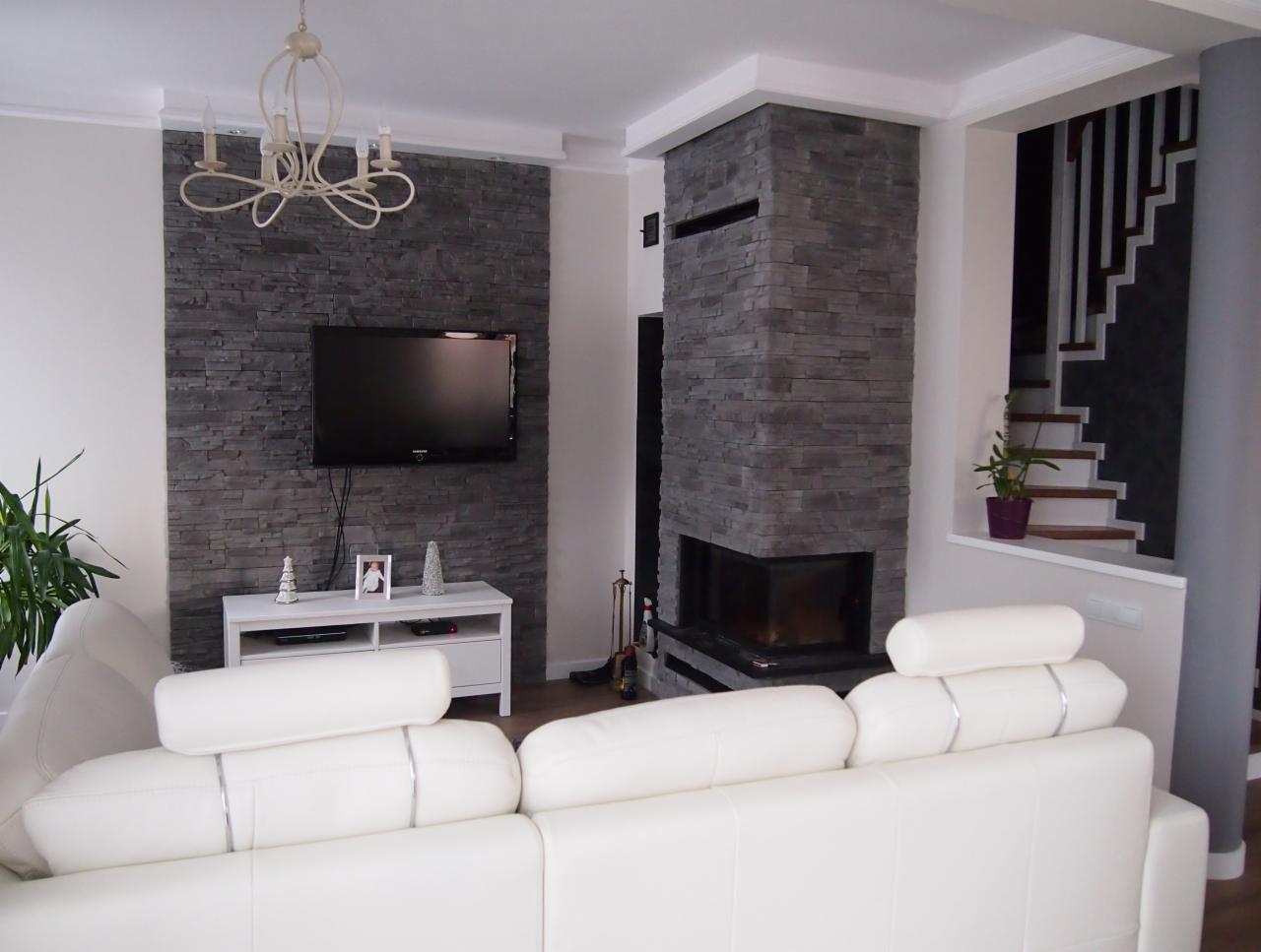 Elegancki salon z kominkiem obłożonym szarym kamieniem, białym narożnikiem i ścianą z telewizorem. Widoczne schody i dekoracyjny żyrandol.