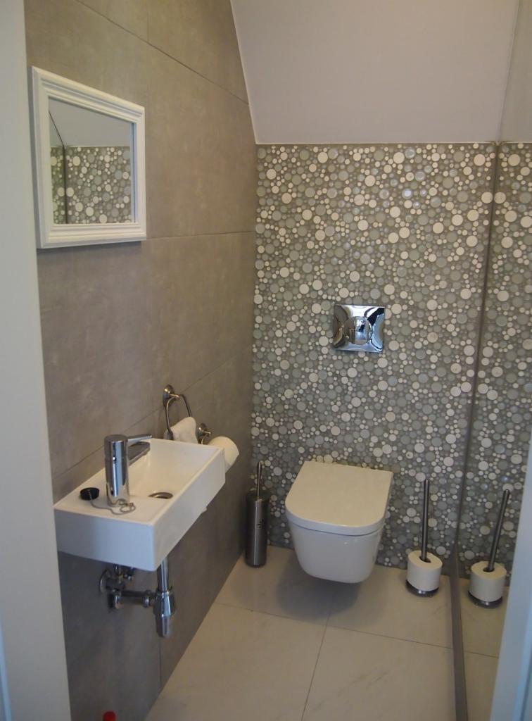 Nowoczesna toaleta w Olsztynie: biała umywalka na szarym tle, lustro i mozaika z kół. Minimalistyczny design, czystość i funkcjonalność.