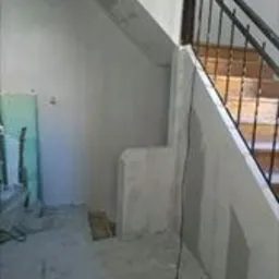 Wnętrze w trakcie prac wykończeniowych: ściany pokryte świeżą warstwą szpachli, widoczne elementy konstrukcji schodów i metalowa balustrada, surowy beton na podłodze, przewody elektryczne zwisające...