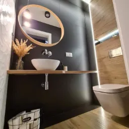 Nowoczesna łazienka z czarnymi płytkami, drewnianym blatem pod umywalką i lustrem w okrągłej ramie, oświetlona taśmami LED, z koszem na papier toaletowy z napisem 'MY STAFF'.